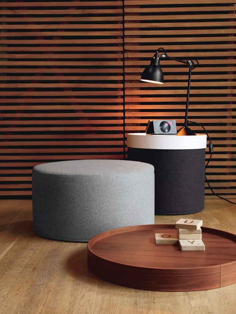 A modern living room featuring a grey ottoman, a round wooden coffee table with wooden blocks on it, and a black and white lamp on a cylindrical table, set against a wooden slatted backdrop.