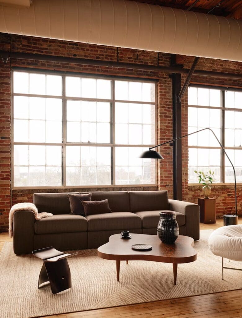 Modern living room featuring a brown leather sofa with decorative pillows, a unique wooden coffee table, and large windows creating a bright, airy atmosphere.