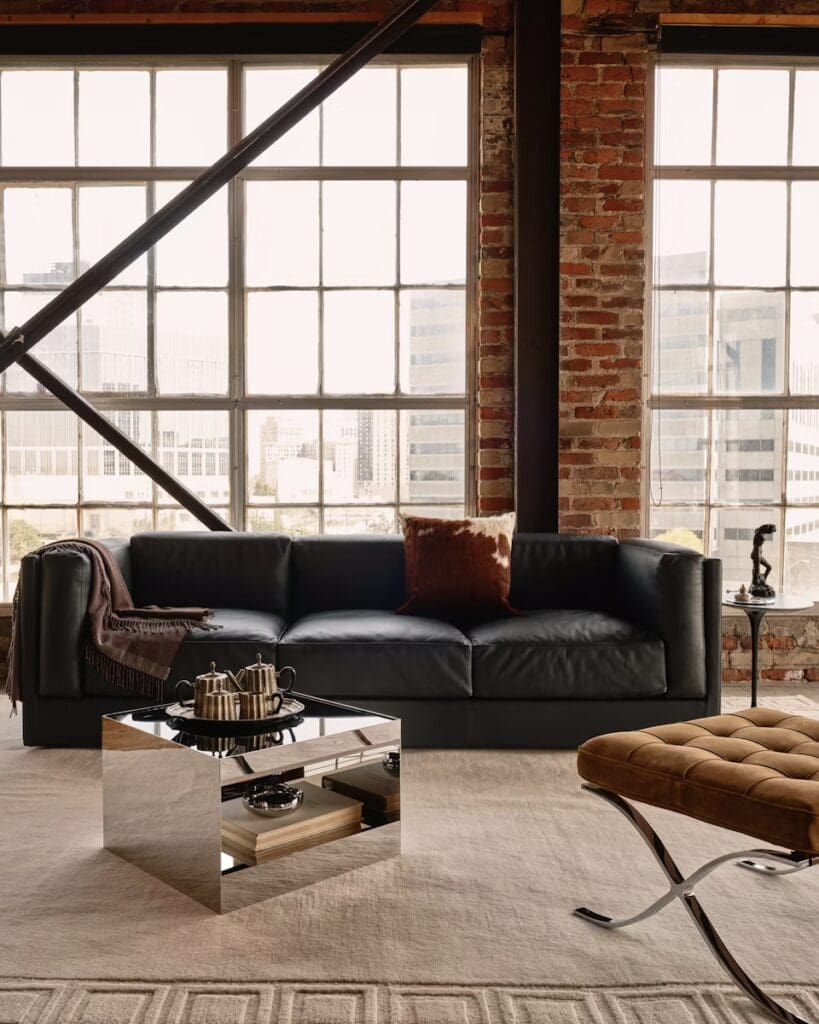 Modern living room featuring a sleek black leather sofa, a mirrored coffee table with elegant serving pieces, and a cozy brown accent pillow, set against a backdrop of large industrial windows.
