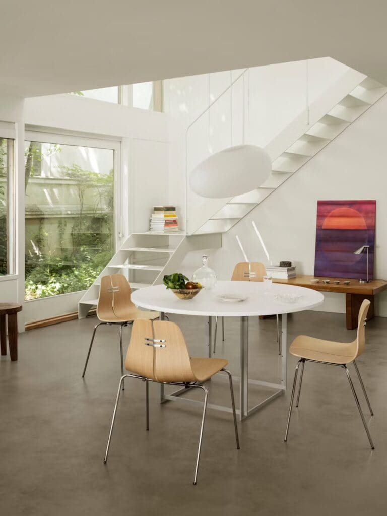 Modern dining area with a round white table, wooden chairs, a glass vase with fruits, and a staircase in a bright space with large windows.