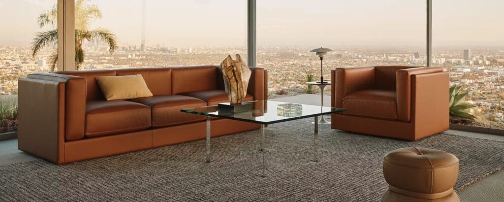Modern living room with a brown leather sofa and armchair, glass coffee table, and a view of the city skyline.