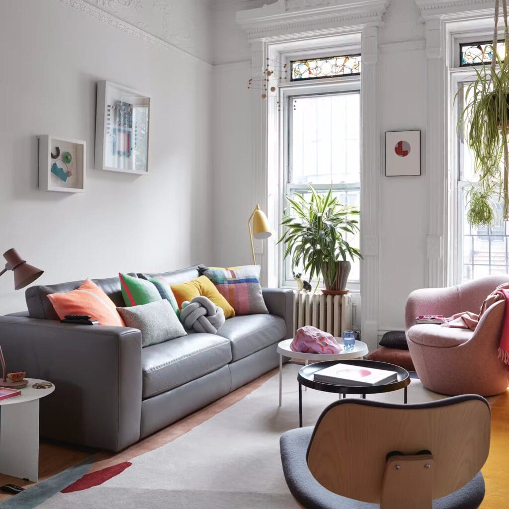 A cozy living room featuring a modern gray sofa adorned with colorful pillows, a round coffee table, and stylish accent chairs. Large windows allow natural light to enter, complemented by greenery from houseplants.