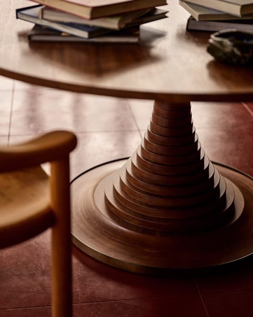 Close-up of a modern wooden table with a spiral base, stacked books on top, and a simple wooden chair in the foreground.