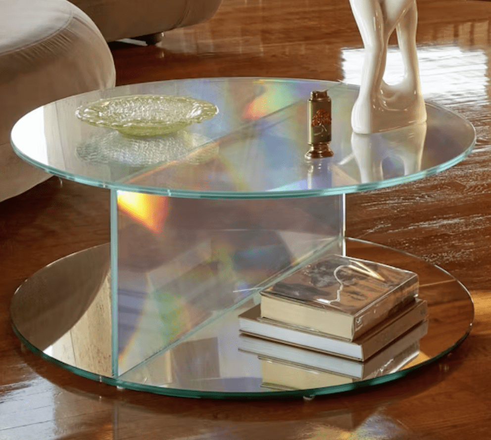 A round glass coffee table with a reflective surface, featuring a decorative dish, a small decorative item, and stacked books underneath.