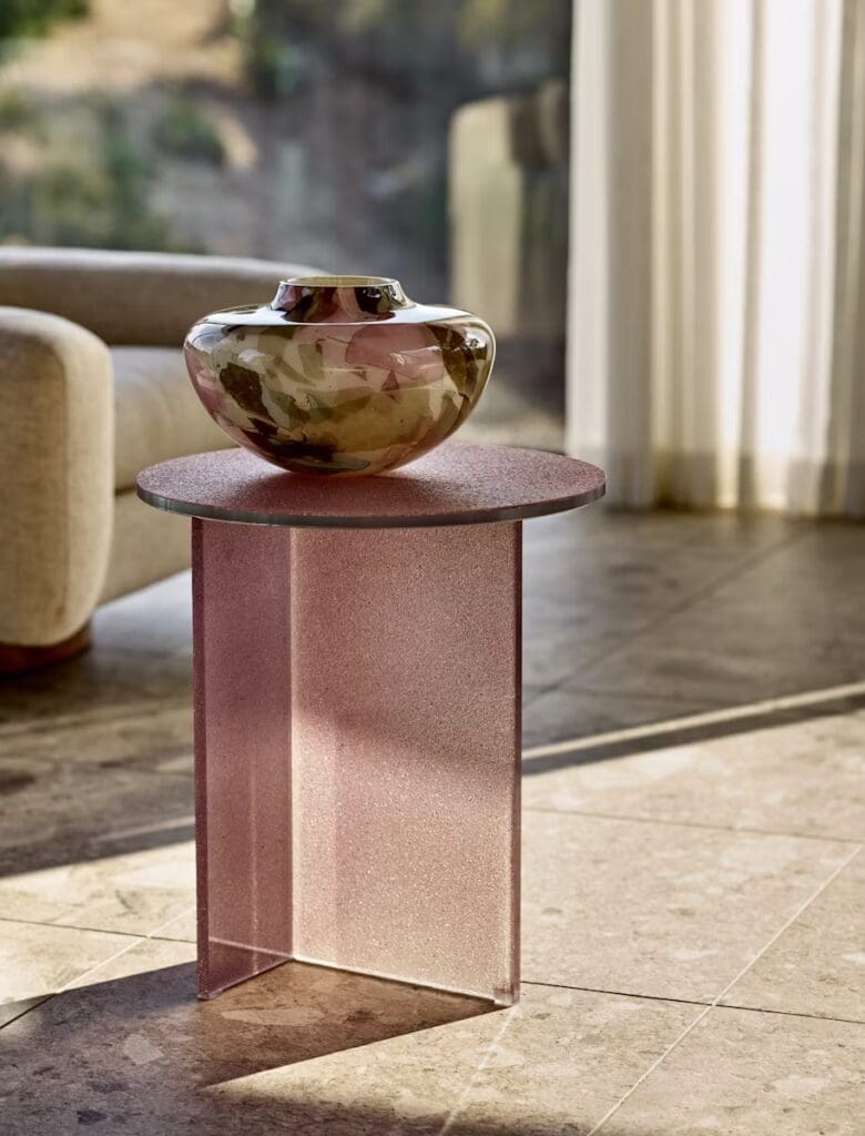 A close-up view of a pink translucent side table with a decorative patterned vase on top, positioned in a modern living room with natural light filtering through sheer curtains.