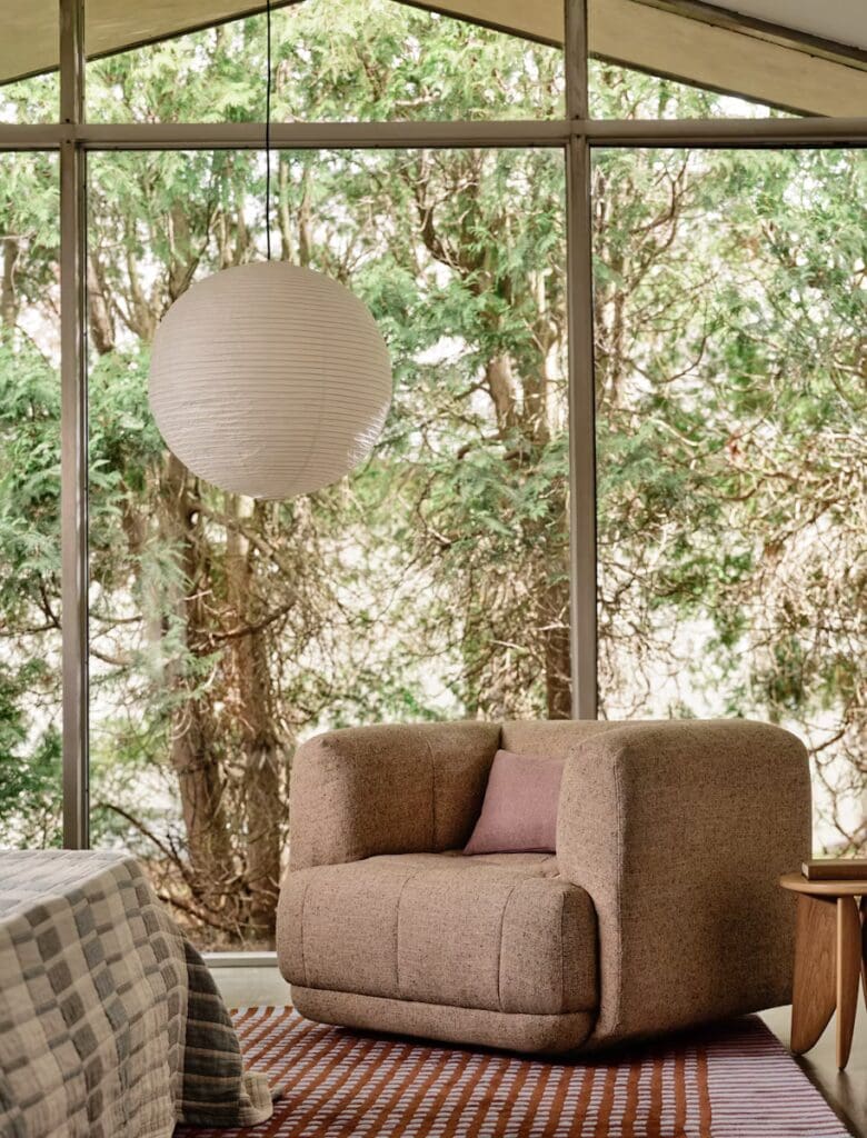 Cozy living room featuring a textured brown armchair with a small pink pillow, complemented by a pendant light and a view of green trees through large windows.