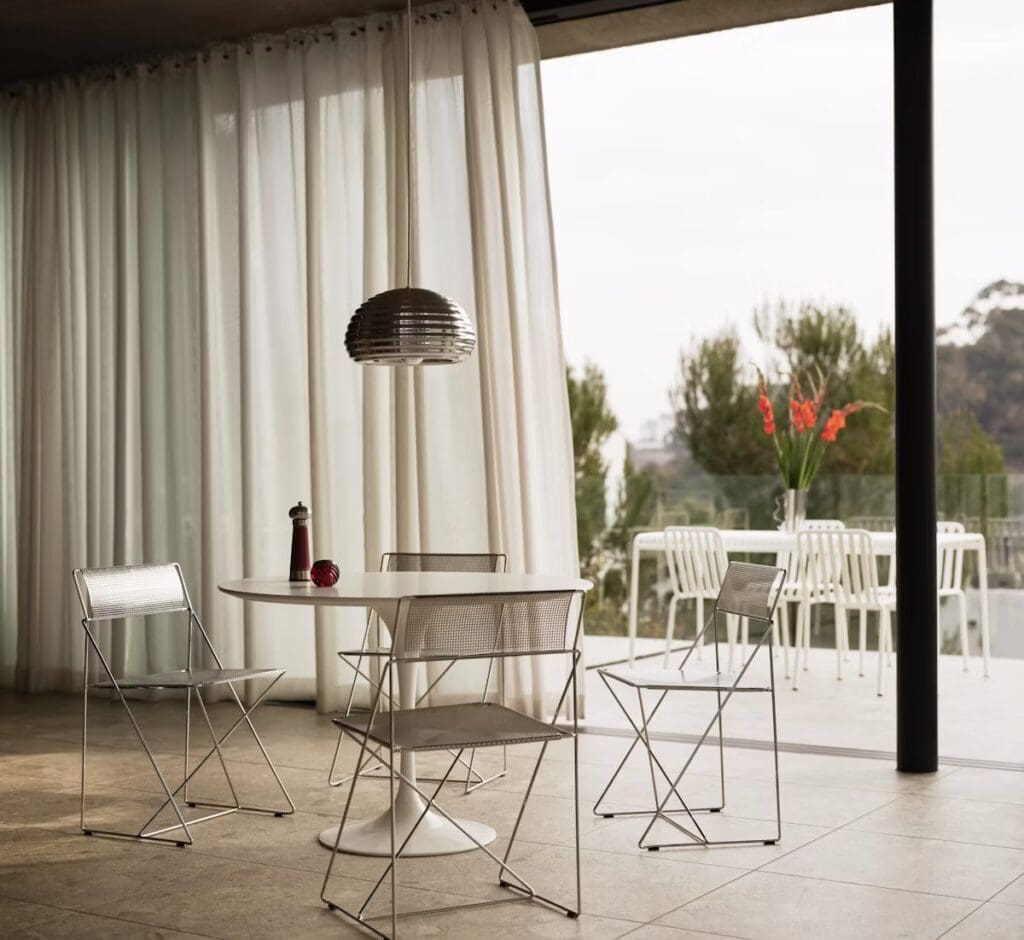 Modern dining area featuring a round white table, four metallic folding chairs, and a pendant light, with large curtains and a view of an outdoor patio.