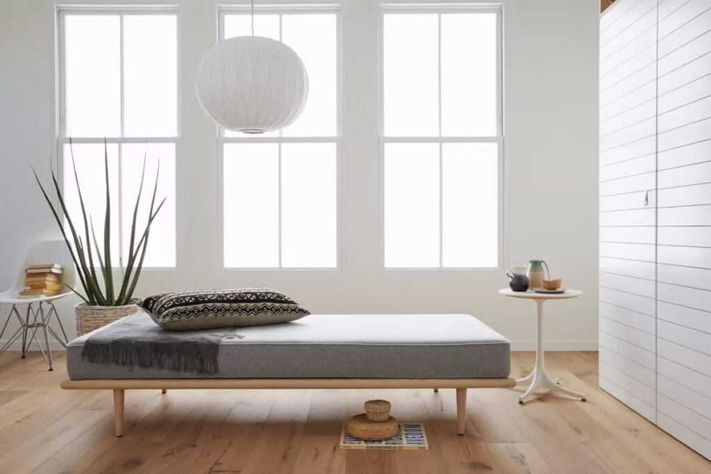 Minimalist living space featuring a low wooden daybed with a gray mattress, decorative black and white pillow, and a cozy throw. A potted plant stands beside the bed, and a small round table holds a carafe and cup. Natural light floods in through large windows.