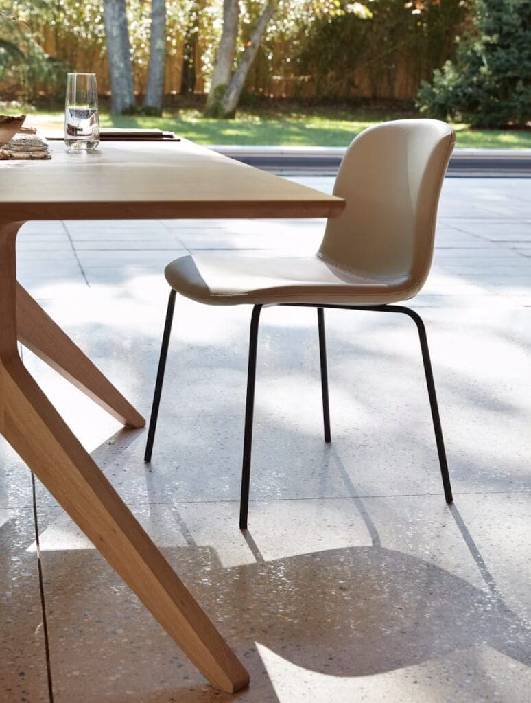 A light-colored dining table with an angled wooden leg and a beige chair nearby, set in a bright interior with a view of a green garden through a large window.