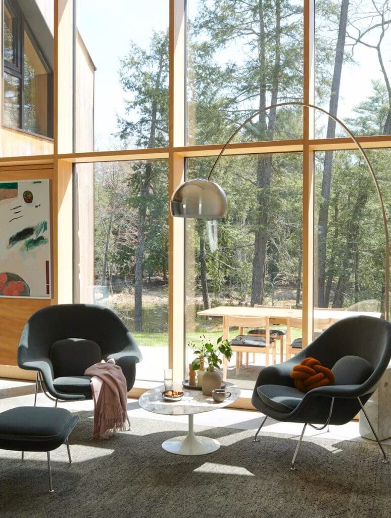 Bright and modern living room featuring two stylish gray chairs, a round marble coffee table, and a contemporary floor lamp, surrounded by large windows that offer a view of the forest outside.