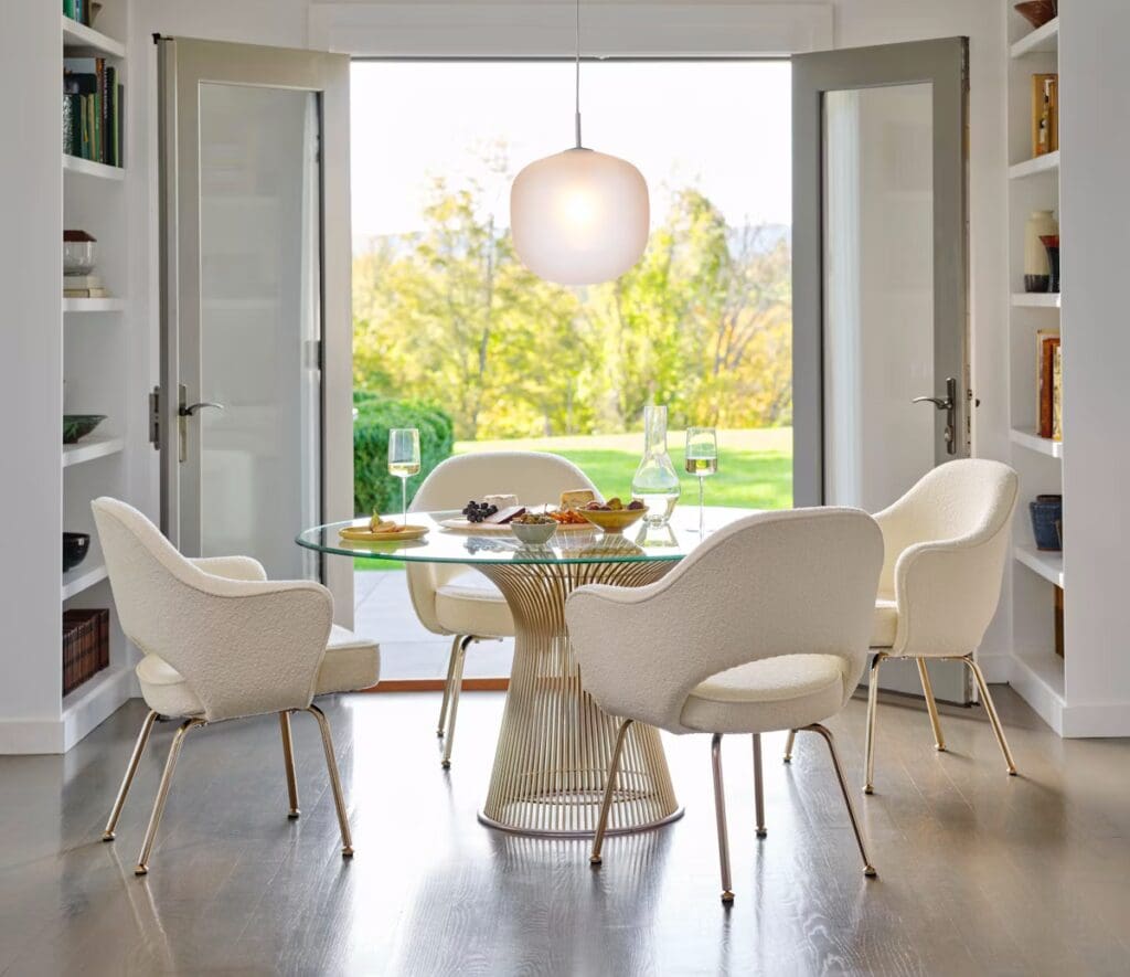 Modern dining area featuring a round glass table surrounded by white upholstered chairs, with snacks and drinks on the table and a view of greenery through open doors.