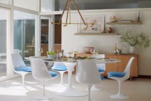 A modern dining room featuring the Knoll Saarinen Oval Dining Table at the center, surrounded by Tulip Chairs with blue cushions. Natural light streams through large windows, highlighting a minimalist decor with a pendant light and decorative shelving.