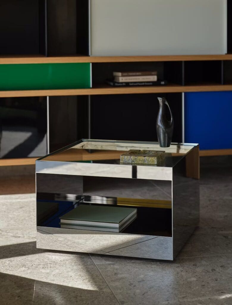 A modern coffee table with a mirrored surface and a green accent shelf in the background, surrounded by books and decorative items.