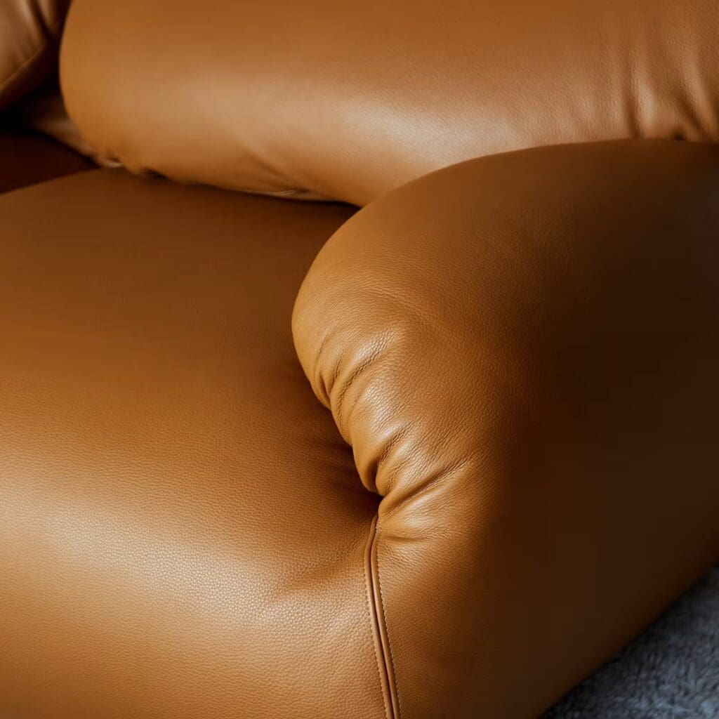 Close-up of a modern leather sofa, featuring a smooth tan upholstery with a rounded armrest and plush cushioning.