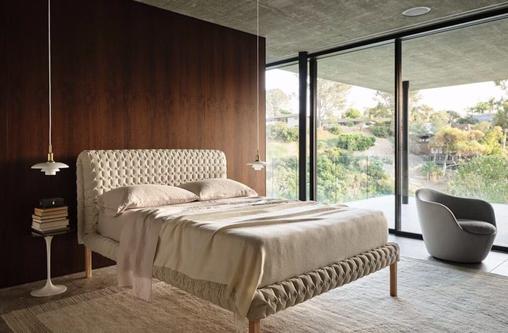 Modern bedroom featuring a tufted bed with beige linens, a bedside lamp, a stack of books, and a cozy armchair next to large glass windows revealing a green landscape outside.