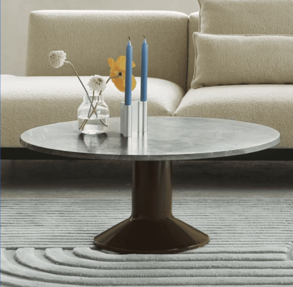 A contemporary living room scene featuring a round marble coffee table with a unique brown base, adorned with a vase of flowers and two blue candles. In the background, a light-colored sofa adds a sophisticated touch.