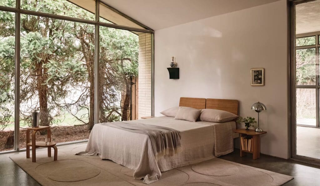 A minimalist bedroom featuring a wooden bed frame with neutral bedding, a small side table with a lamp, and a large window showcasing greenery outside.
