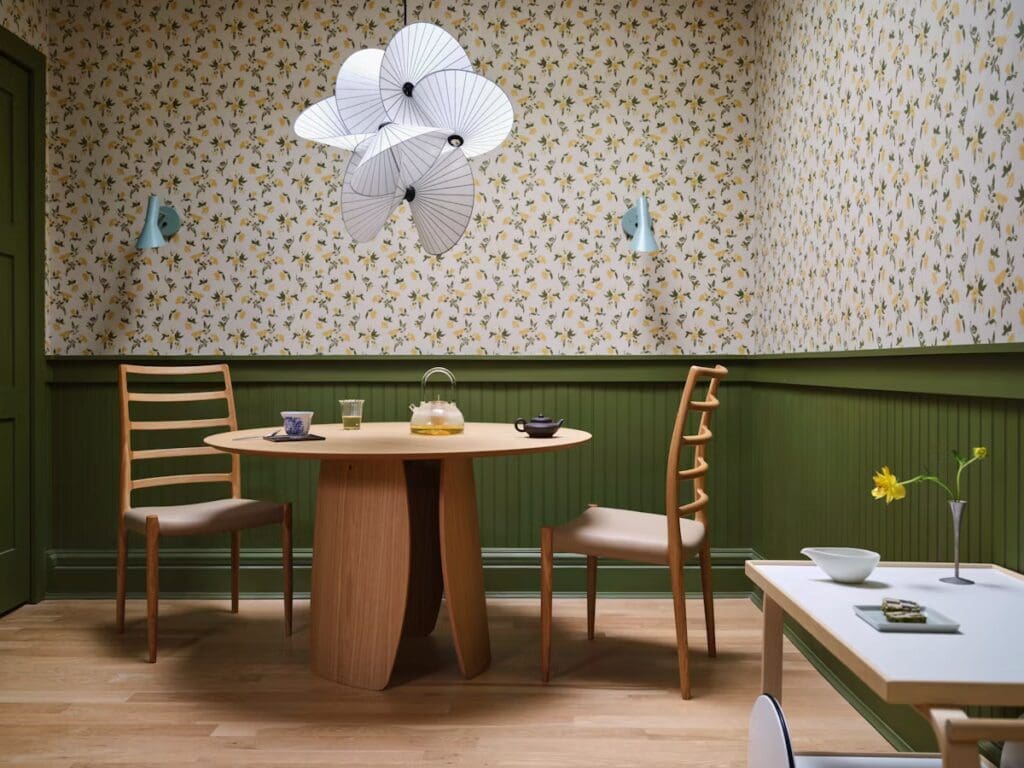 Cozy dining area featuring a round wooden table, two wooden chairs, and a floral wallpaper design. A stylish pendant light hangs above, and various tea items are arranged on the table.