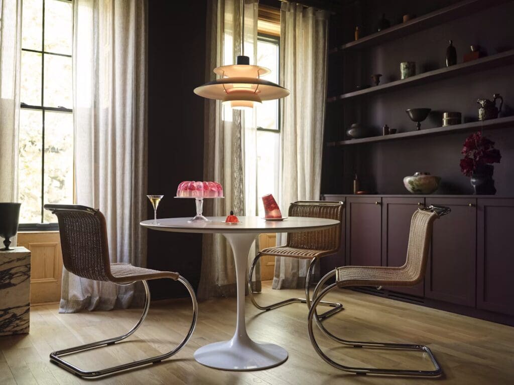 A dining area featuring Eero Saarinen's Tulip Table, surrounded by two woven chairs with chrome bases. The space is illuminated by a modern pendant light and decorated with various objects on a dark shelving unit and a window with sheer curtains.