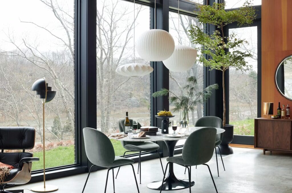 Modern dining room featuring the Knoll Saarinen Oval Dining Table with Tulip Chairs, large windows showcasing a natural landscape, stylish pendant lighting, and contemporary decor.