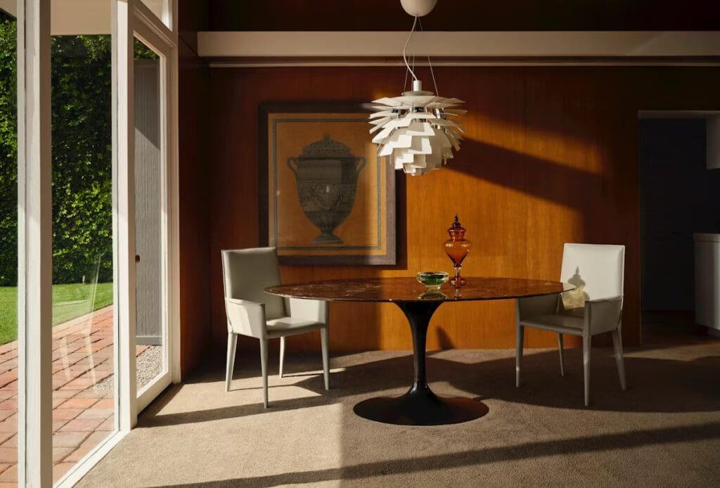 A stylish dining room featuring a Knoll Saarinen Oval Dining Table with a dark, glossy surface and a Tulip Chair on either side, illuminated by a unique pendant light. A decorative vase sits on the table, with a large artwork on the wall behind it and sunlight streaming in through large windows.