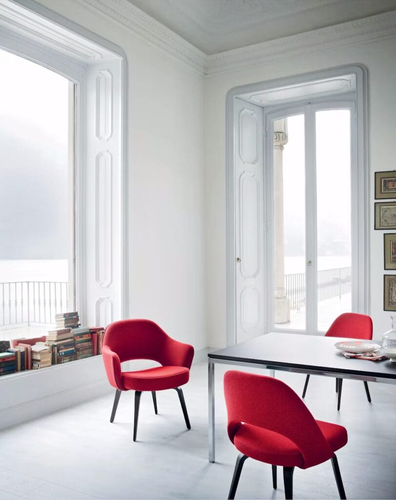 A modern interior featuring a dining table and two red chairs with sleek black legs, set against large windows and a bookshelf filled with books.