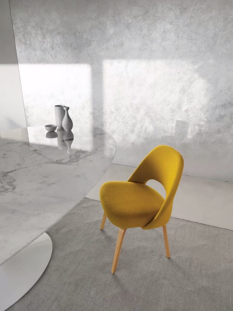 A modern yellow chair with wooden legs placed beside a round marble table, with minimalist vases on the table and a textured light gray wall in the background.