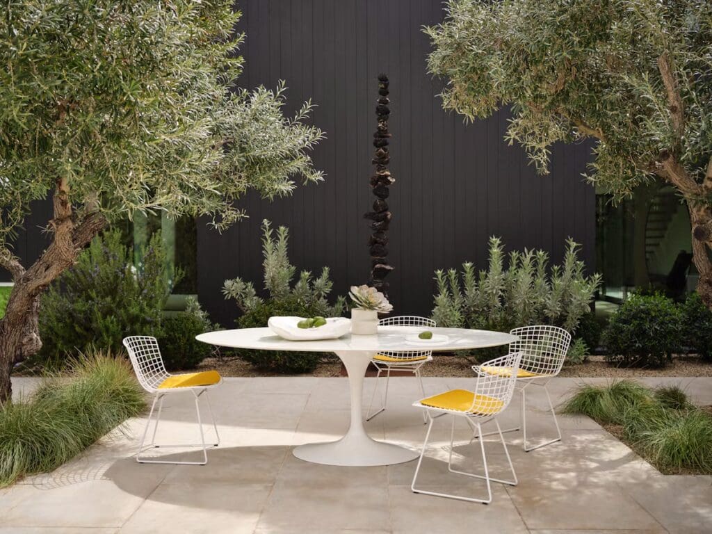 A modern outdoor dining setup featuring a round white table with a central centerpiece, surrounded by four wire chairs with yellow cushions, set against a backdrop of olive trees and a minimalist architectural wall.