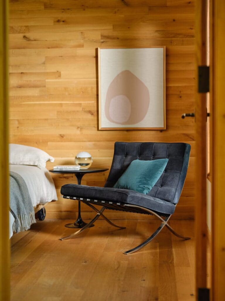 A stylish chair with a dark fabric and a green cushion sits beside a small side table with a decorative globe. In the background, there is a bed with white bedding and a wooden wall with a framed abstract art piece.