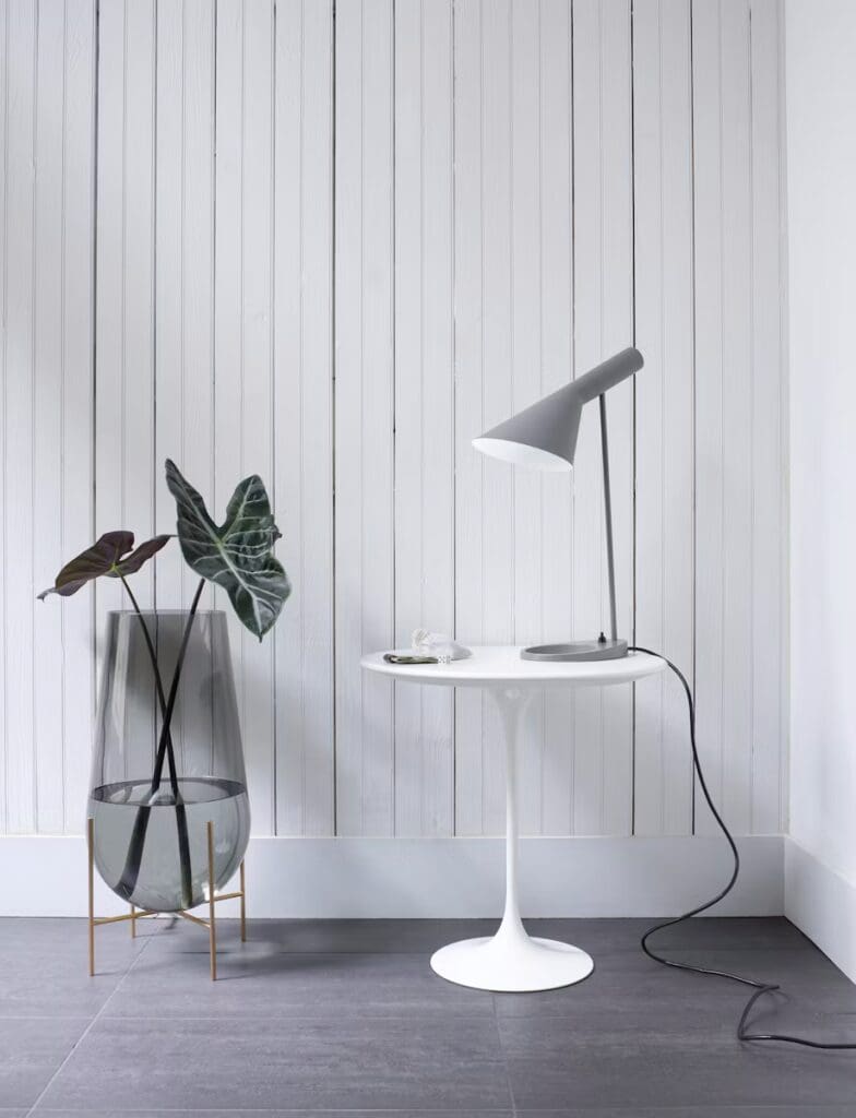 A contemporary interior featuring a white tulip side table with a gray lamp on top, accompanied by a glass planter holding tall dark green plants, set against a backdrop of vertical white wood paneling.