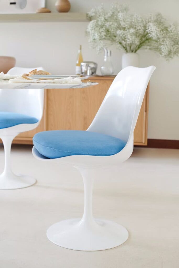 A modern Tulip™ chair designed by Eero Saarinen, featuring a sleek white base and a blue cushion, placed at a dining table with food in the foreground.