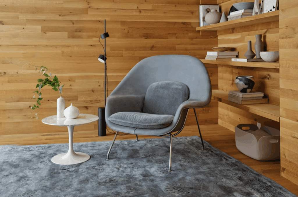 A modern living room featuring a gray Womb Chair by Eero Saarinen, a white side table with a decorative vase and plant, and wooden shelves displaying various objects against a warm wood wall.