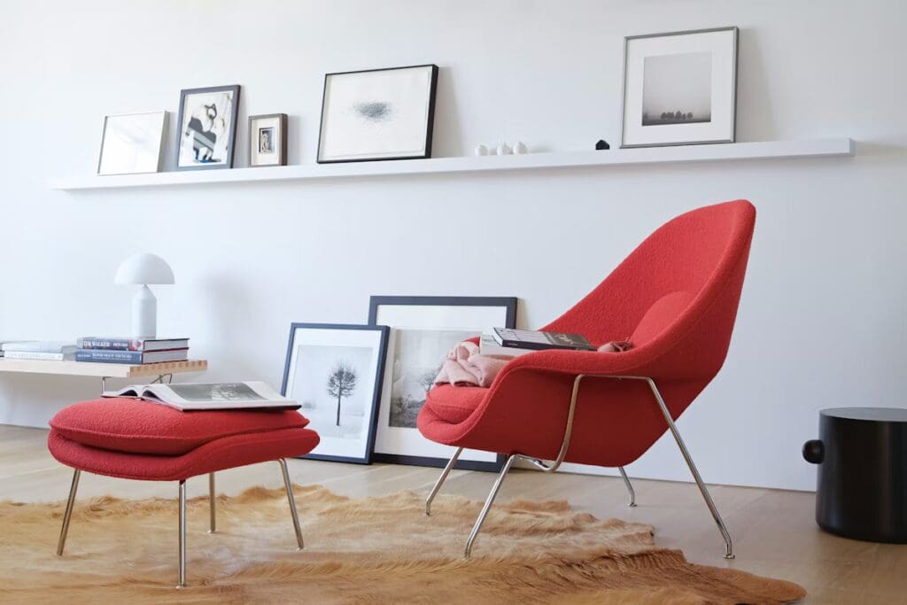 A modern living space featuring a red Womb Chair and ottoman by Eero Saarinen, with a simple shelf displaying framed art and a stylish lamp in the background.