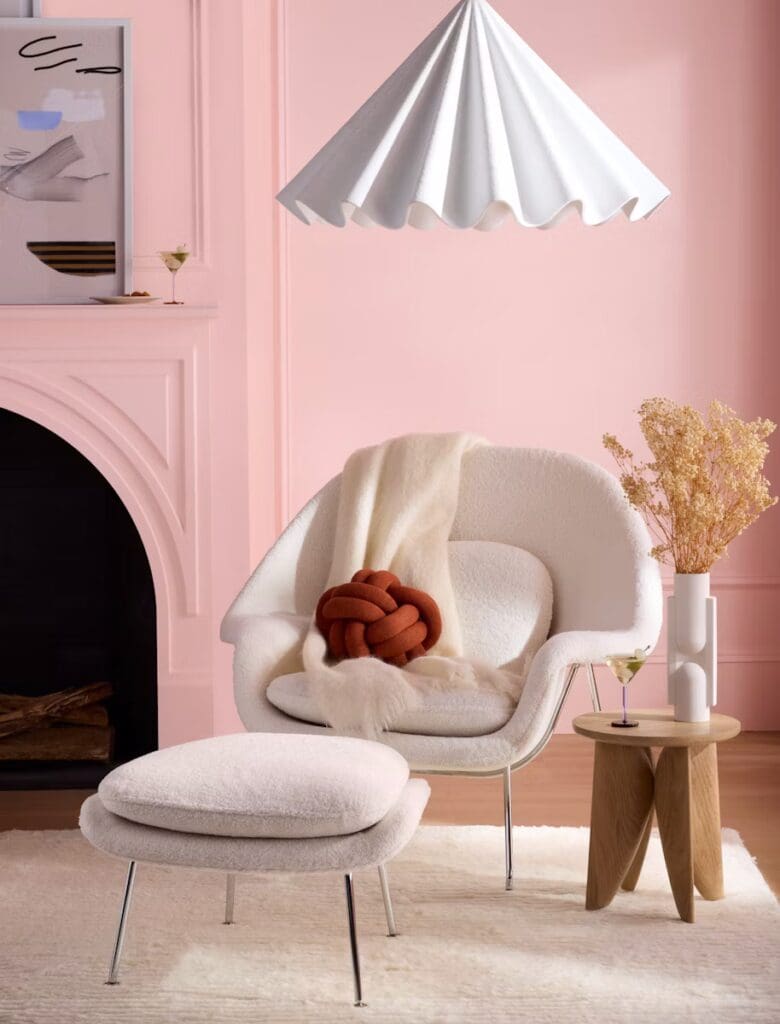 A cozy living space featuring a white Womb™ Chair and ottoman, adorned with a textured pillow, a throw blanket, and a nearby wooden side table. The room has pastel pink walls and a unique conical lamp hanging above.